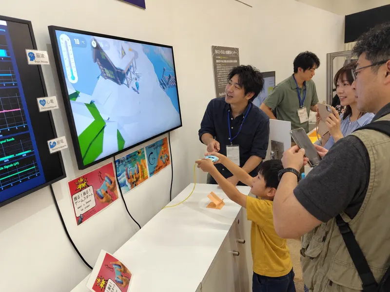 At the Expo booth, a child plays with the experience device in front of the game screen while a staff member explains and other visitors look on